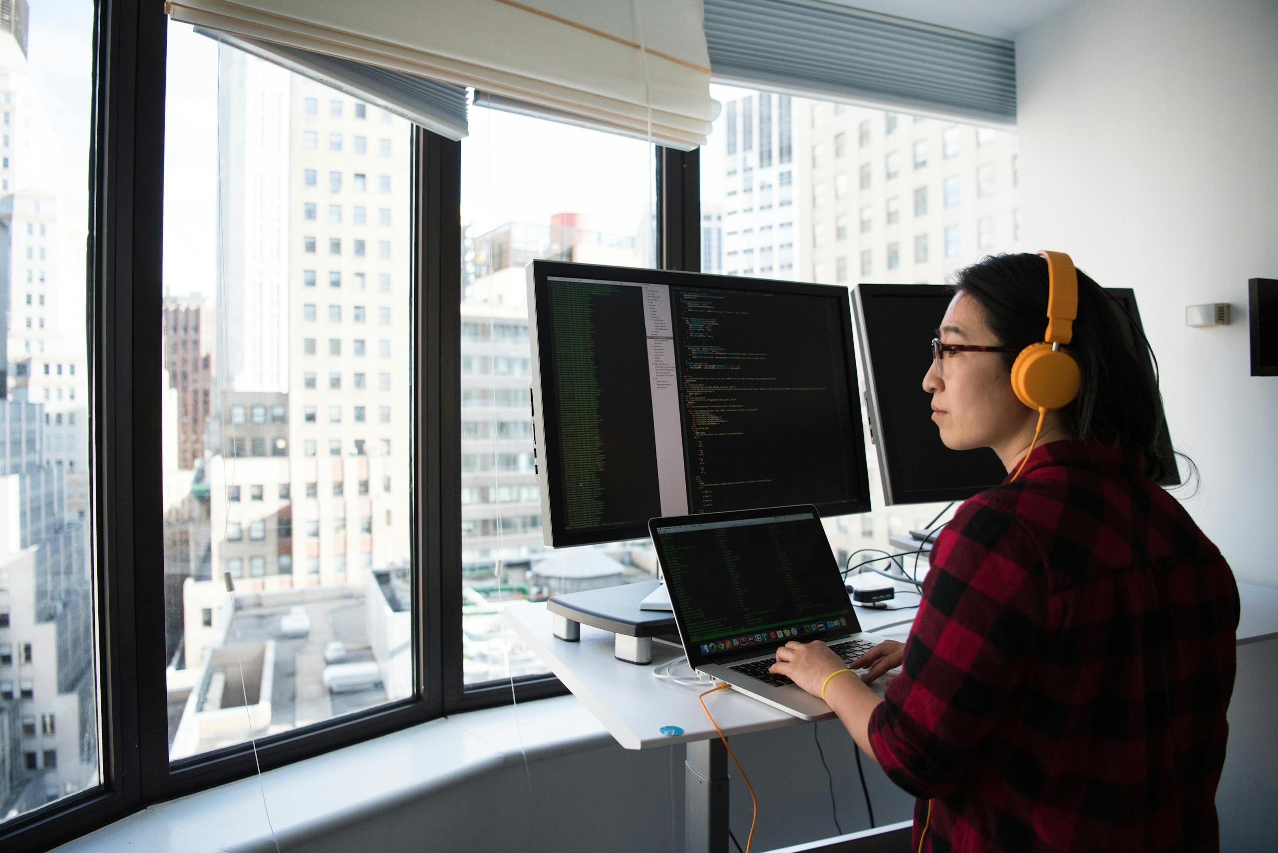 A businesswoman coding at a standing desk with city views through large windows, wearing headphones.